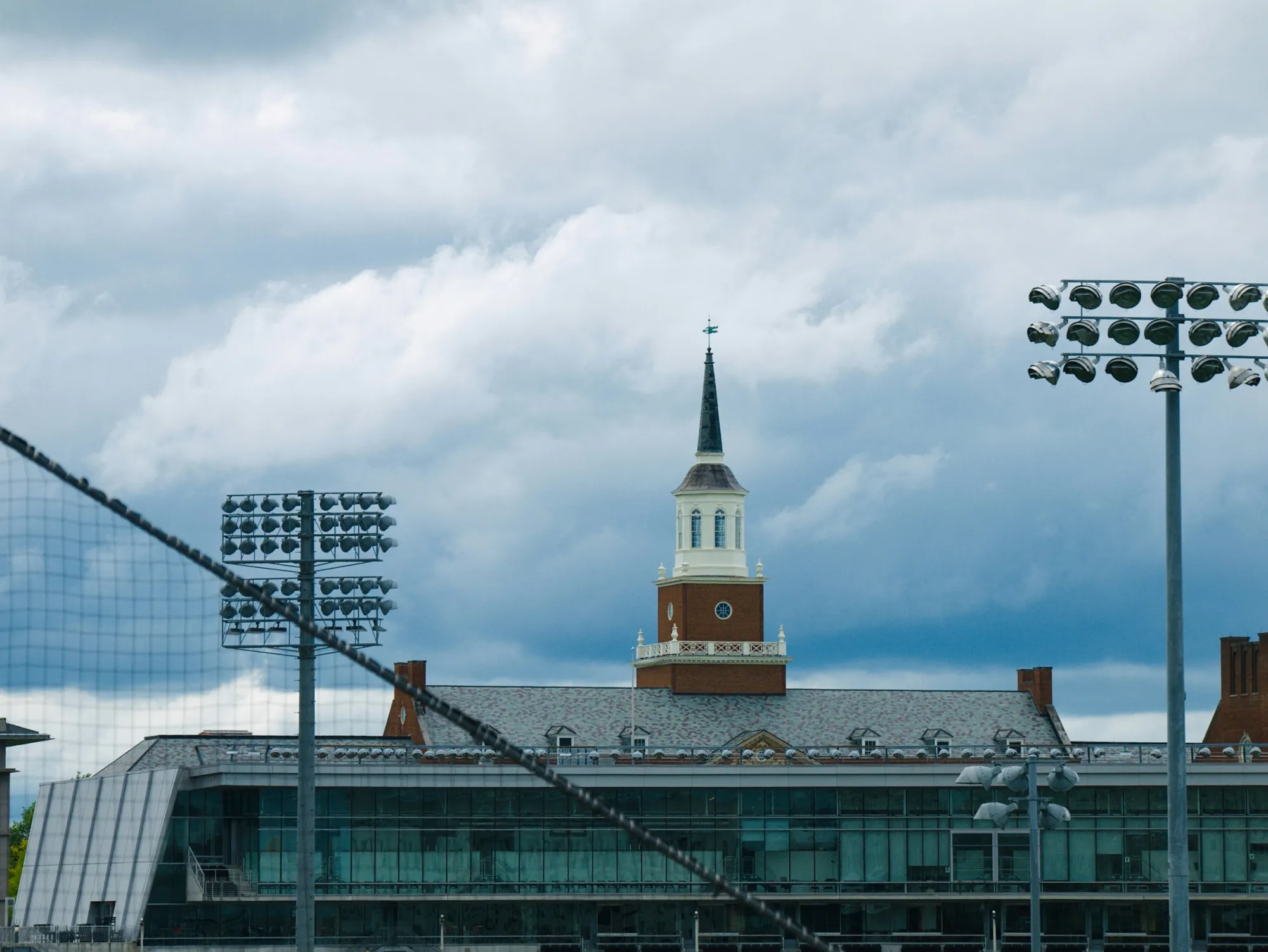 Brick university building with a white clock tower and steeple behind stadium floodlights under a cloudy sky