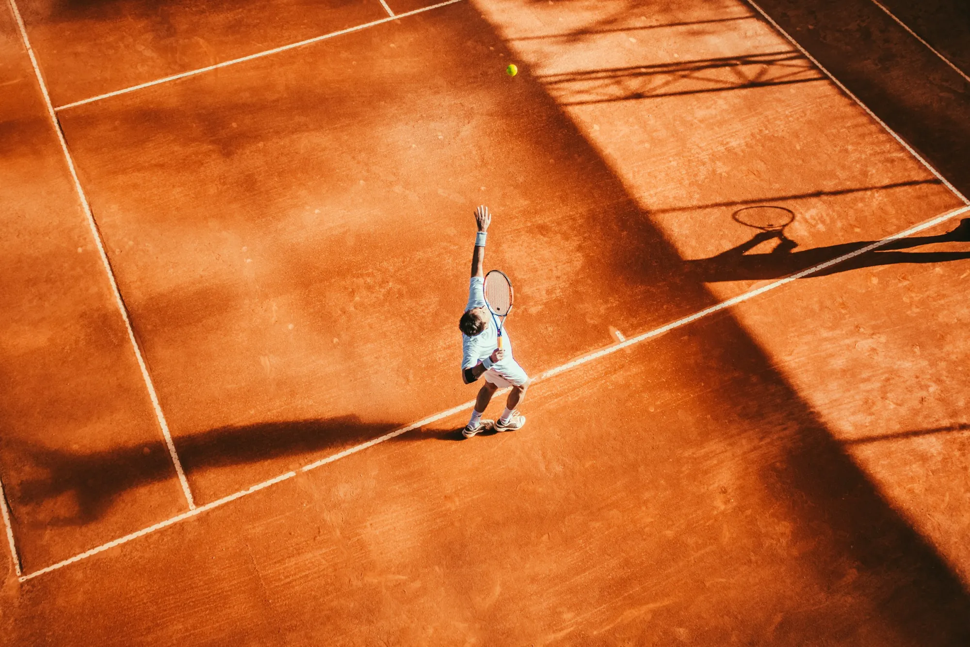 Tennis player serving on a clay court, seen from above in late afternoon light