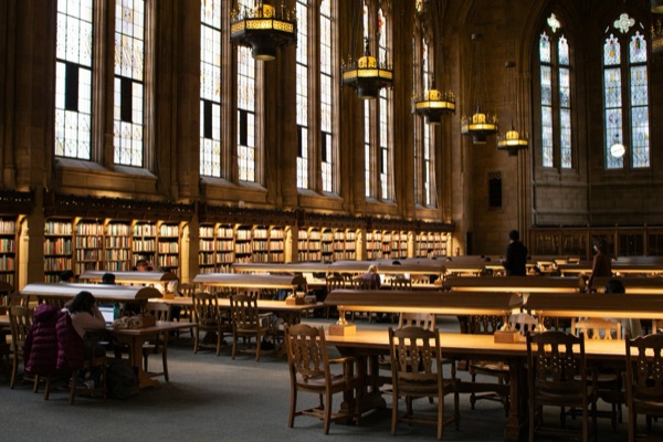 Students studying at long wooden tables in a historic university library reading room with tall stained glass windows