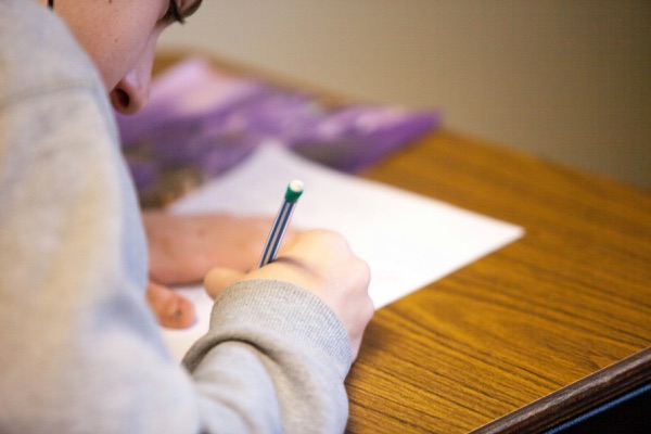 Close-up of a student writing with a pencil on a sheet of paper at a wooden desk