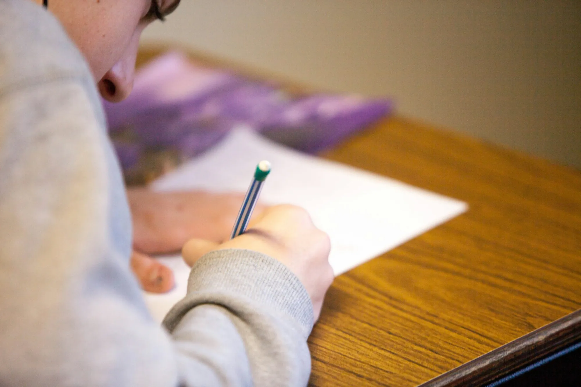 Primer plano de un estudiante escribiendo con un lápiz sobre una hoja en un pupitre de madera