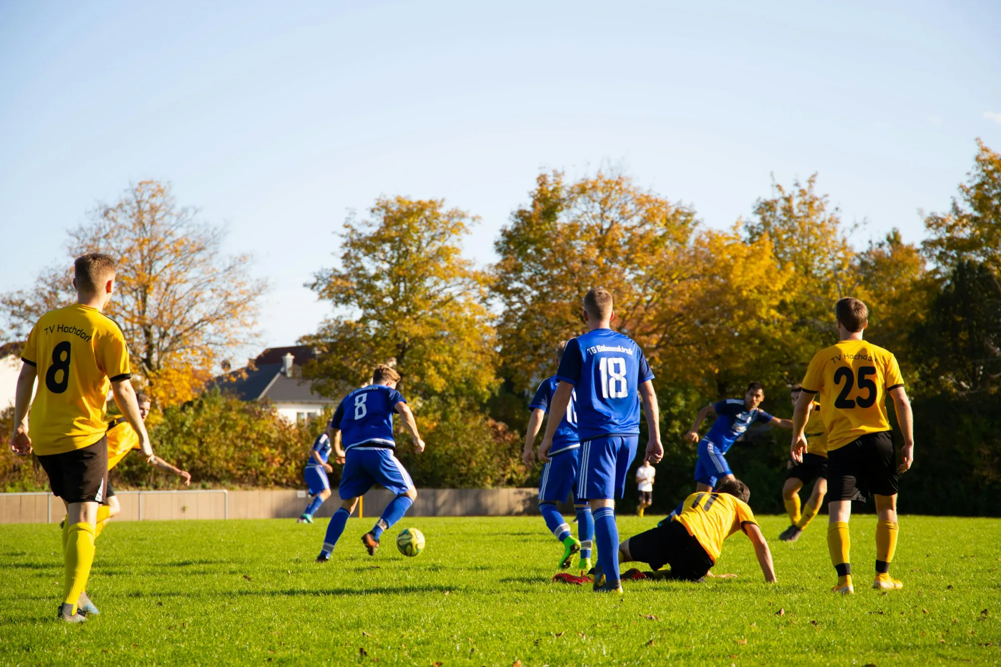 Dos equipos de fútbol con camisetas azules y amarillas jugando en un campo de césped un día soleado de otoño