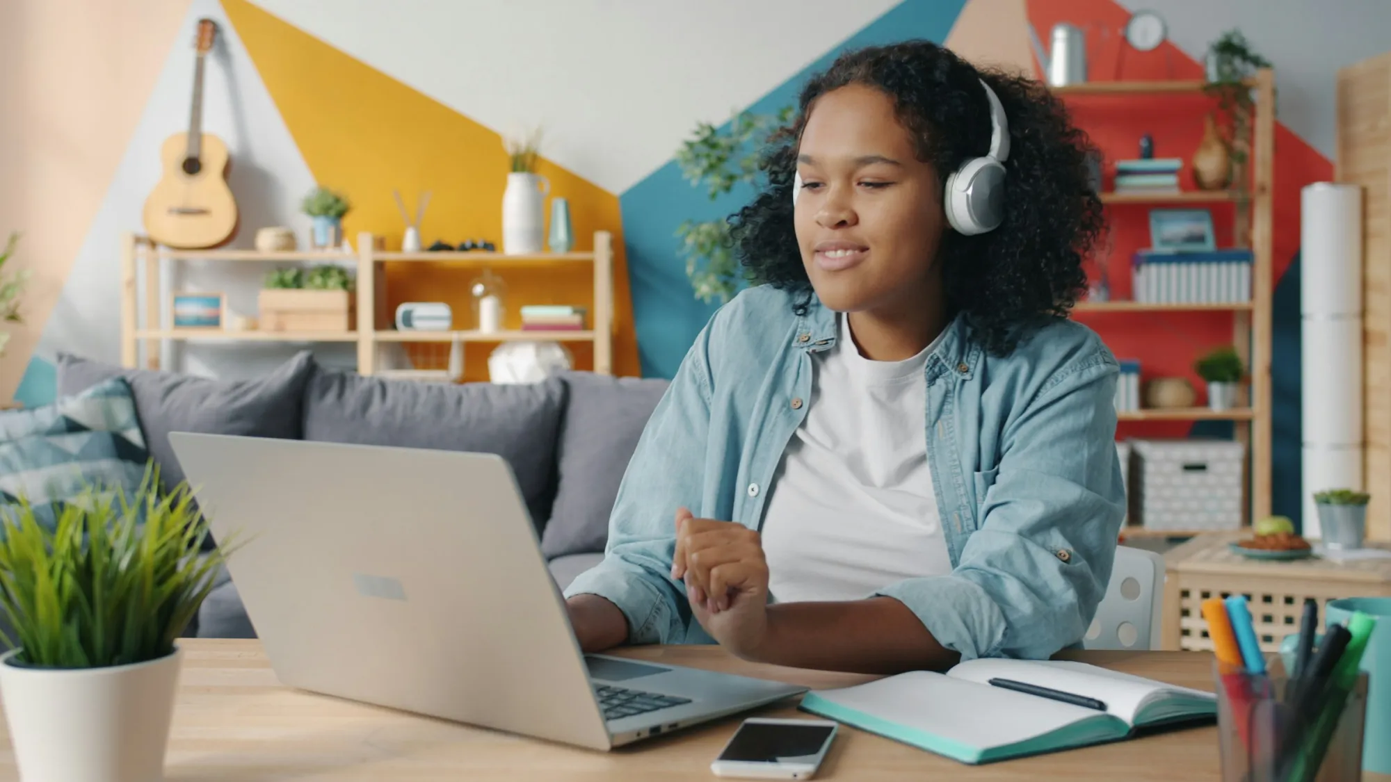 Young woman with headphones working on a laptop at a desk at home, with a notebook next to her